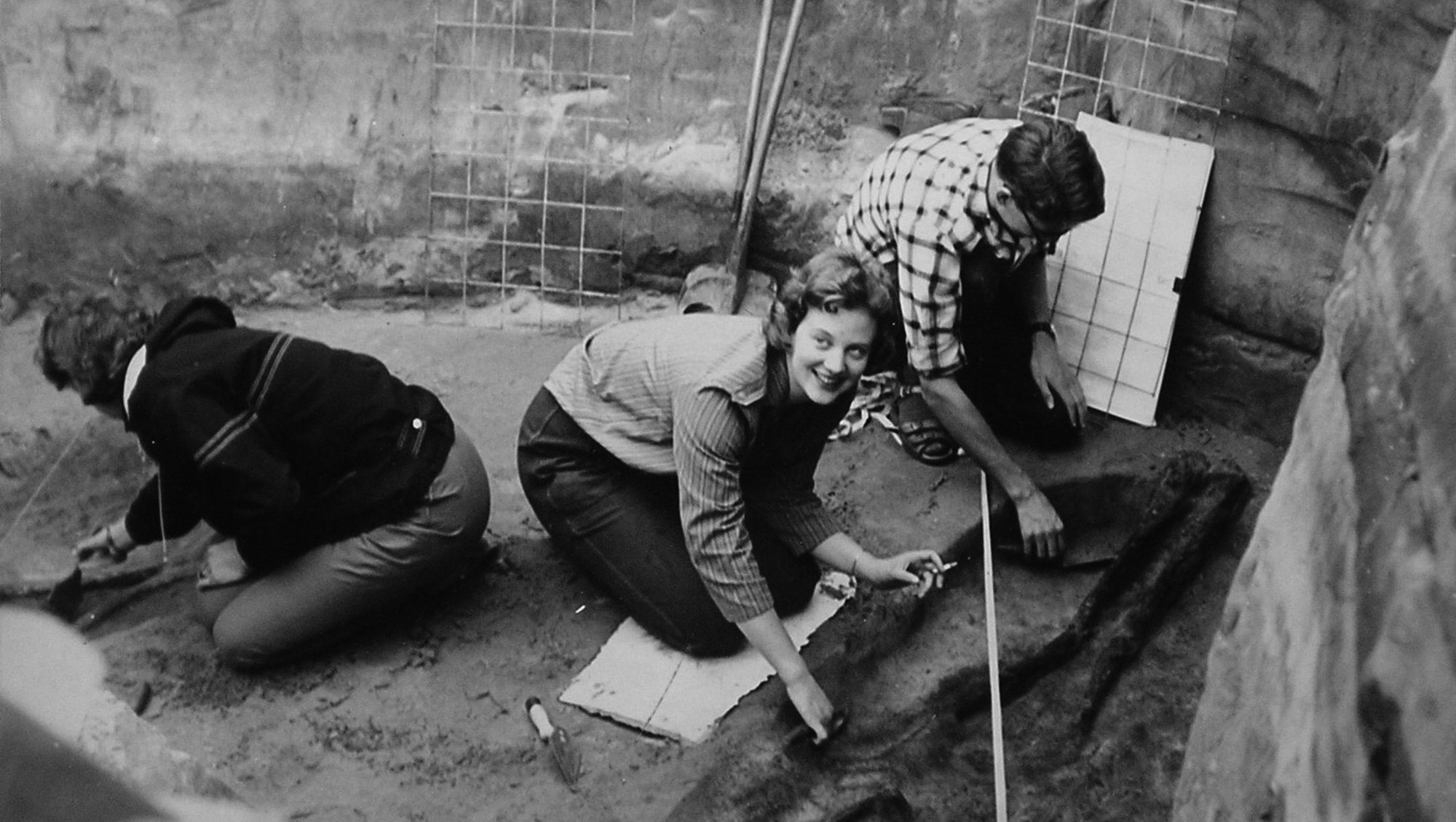 Prinsesse Margrethe sidder i en udgravning sammen med 2 andre personer og kigger op, mens de 2 andre kigger ned og er i gang med at udgrave.