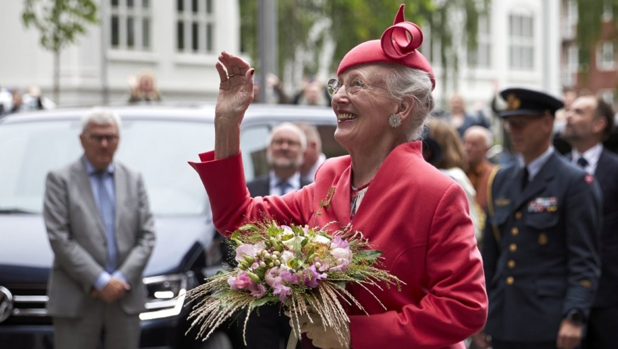 Dronning Margrethe iført en pink dragt og hat og med en stor buket blomster, vinker og smiler.