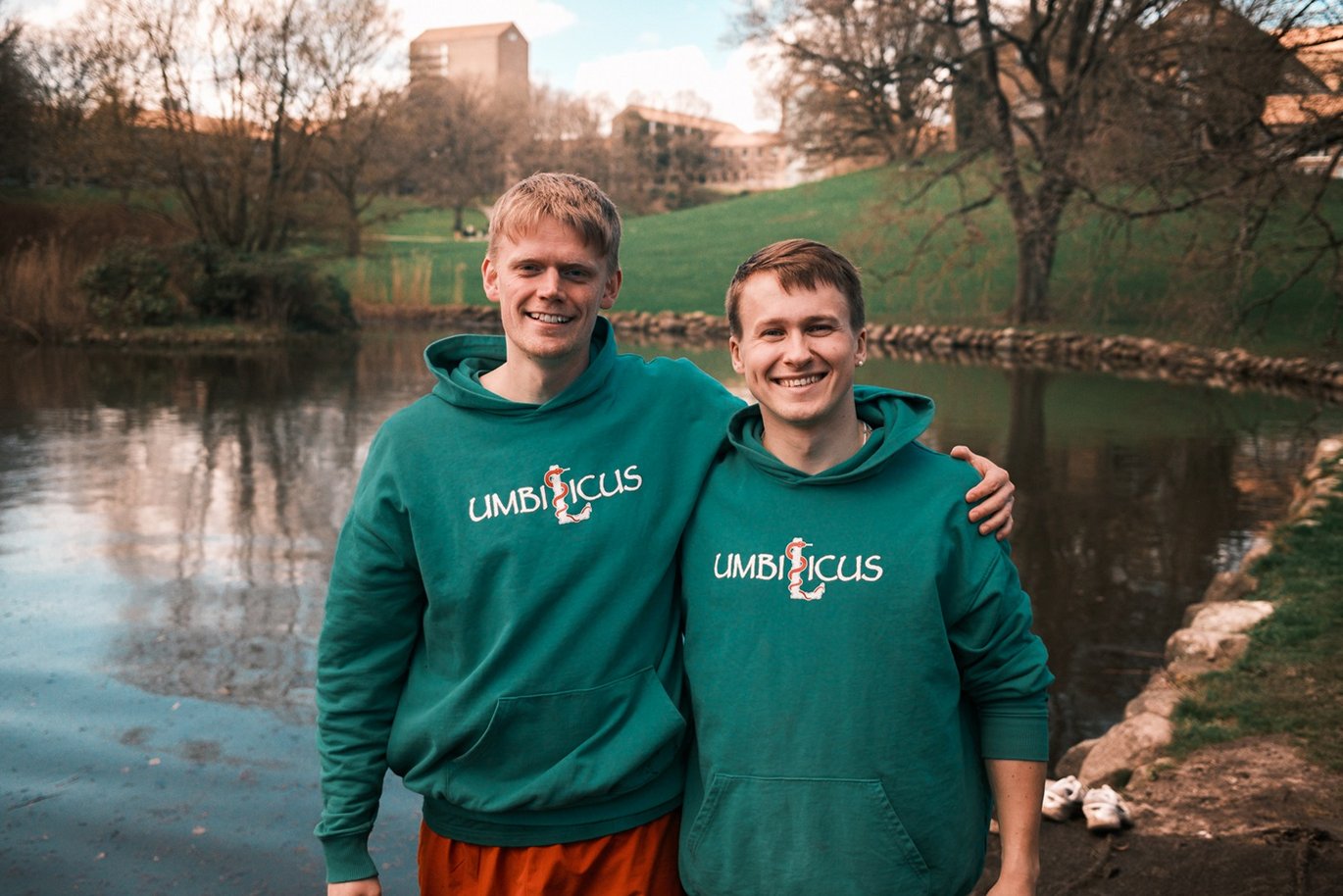 Daniel Teglborg Houe og Morten Wittendorf Bækby, begge iført en Umbilicus-hættetrøje, står foran søen i universitetsparken.
