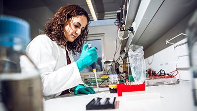 Kvinde i hvid laboratoriekittel iført handsker sidder ved et bord med en pipette.
